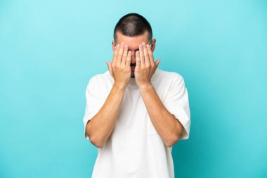 Genç beyaz adam mavi arka planda izole edildi. Gözlerini elleriyle kapadı.