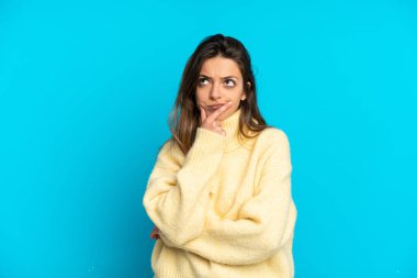 Kafkasyalı genç bir kadın mavi arka planda izole edilmiş, yukarı bakarken şüpheleri var.