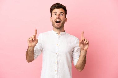 Pembe arka planda izole edilmiş beyaz, yakışıklı genç bir adam şaşırmış ve yukarı bakıyor.