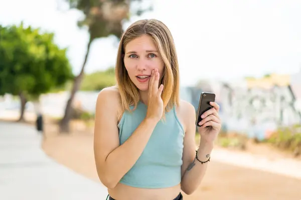 Young Blonde Woman Using Mobile Phone Outdoors Whispering Something — Stock Photo, Image
