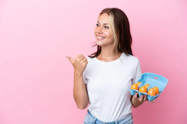 年轻的俄罗斯女人拿着用粉色背景分离出来的卵子 指着旁边展示一种产品 — 图库照片