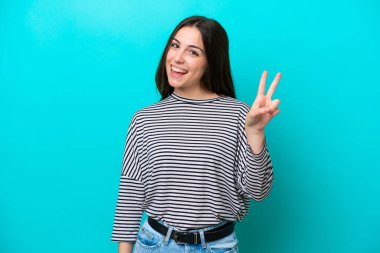 Mavi arka planda izole edilmiş genç beyaz kadın gülümsüyor ve zafer işareti gösteriyor.