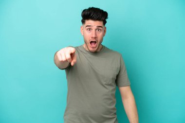 Mavi arka planda izole edilmiş genç beyaz adam hayal kırıklığına uğramış ve ön tarafı işaret ediyor.