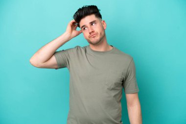 Kafkasyalı genç adam mavi arka planda izole edilmiş, hayal kırıklığı ifadesi var ve anlamıyor.