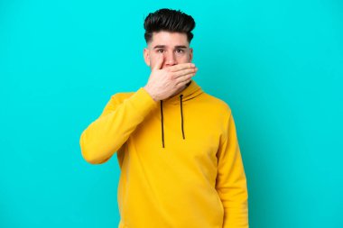 Genç beyaz adam mavi arka planda izole edilmiş, ağzı elle kapatılmış.