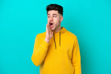 Mavi arka planda izole edilmiş genç beyaz adam yan tarafa bakarken sürpriz bir hareketle bir şeyler fısıldıyor.