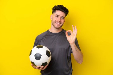 Sarı arka planda izole edilmiş yakışıklı genç futbolcu parmaklarıyla tamam işareti gösteriyor