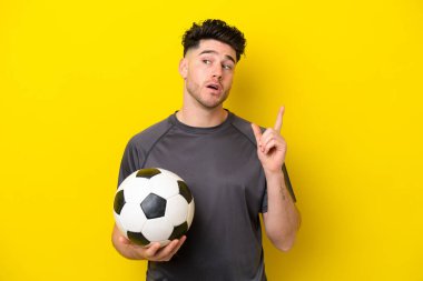Yakışıklı genç futbolcu sarı arka planda izole edilmiş bir şekilde parmağını kaldıran bir fikir düşünüyor.