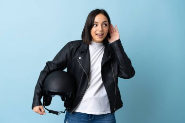 Mavi arka planda izole edilmiş motosiklet kaskı tutan bir kadın kulağına elini koyarak bir şeyler dinliyor.
