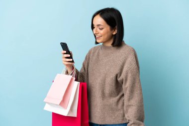 Kafkasyalı genç bir kadın mavi arka planda izole edilmiş. Elinde alışveriş poşetleri var ve bir arkadaşına cep telefonuyla bir mesaj yazıyor.