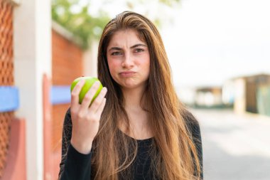 Genç, güzel, beyaz bir kadın, dışarıda elmalı, üzgün bir ifadeyle.