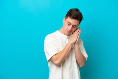 Genç ve yakışıklı Brezilyalı bir adam mavi arka planda izole edilmiş bir şekilde uyku jesti yapıyor.