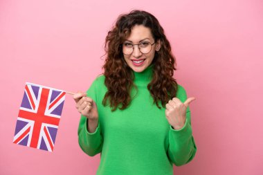 Pembe arka planda İngiliz bayrağı taşıyan beyaz bir kadın bir ürünü sunmak için kenarı işaret ediyor.