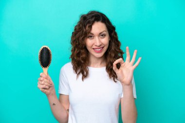 Mavi arka planda saç fırçası tutan genç beyaz kadın parmaklarıyla tamam işareti gösteriyor.