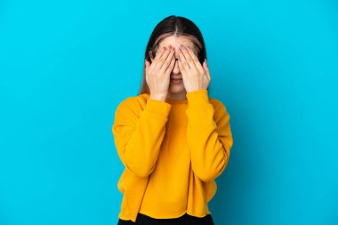 Mavi arka planda izole edilmiş genç beyaz kadın gözlerini elleriyle kapatıyor.