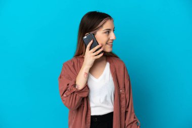 Mavi arka planda izole edilmiş genç beyaz bir kadın cep telefonuyla sohbet ediyor.