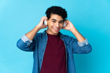 Genç Venezuelalı adam mavi arka planda izole edilmiş müzik dinliyor.