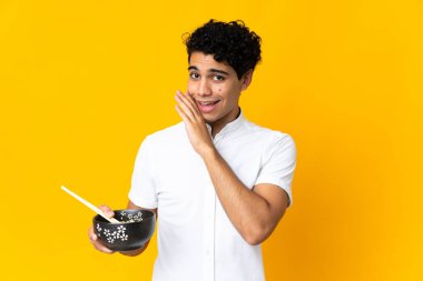 Genç Venezuelalı bir adam sarı arka planda izole edilmiş yemek çubuklarıyla bir kase erişte tutarken bir şeyler fısıldıyor.