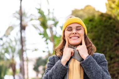 Açık havada kışlık ceket giyen esmer kadın mutlu ve hoş bir ifadeyle gülümsüyor.