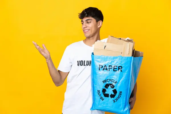 孤立した黄色の背景の上にリサイクルするために紙でいっぱいのリサイクルバッグを持っている男が来るように招待するために手を伸ばします — ストック写真