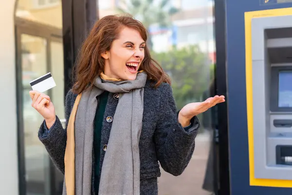 Brunette woman holding a credit card at outdoors with surprise facial expression