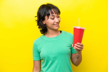 Arjantinli genç bir kadın sarı arka planda izole edilmiş bir gazoz tutuyor. Yan tarafa bakıyor ve gülümsüyor.