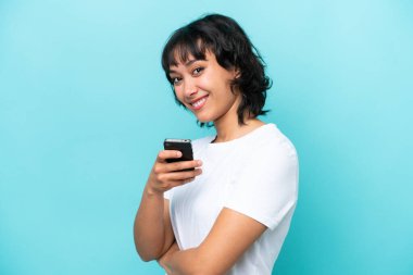 Arjantinli genç bir kadın mavi arka planda izole edilmiş cep telefonu tutuyor ve kollarını kavuşturuyor.