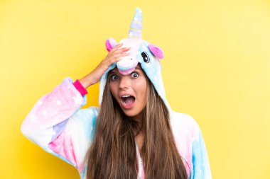 Sarı arka planda izole edilmiş tek boynuzlu at pijaması giyen beyaz bir kadın yan tarafa bakarken sürpriz jest yapıyor.