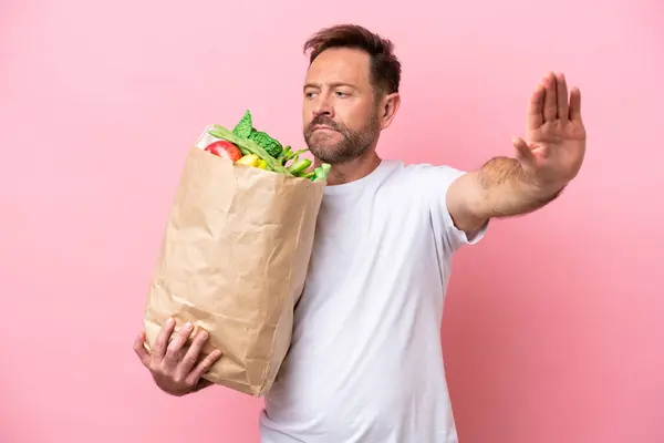 ピンクのバックグラウンドで隔離された食料品のショッピングバッグを保持している中年男性は 停止ジェスチャーと失望 — ストック写真