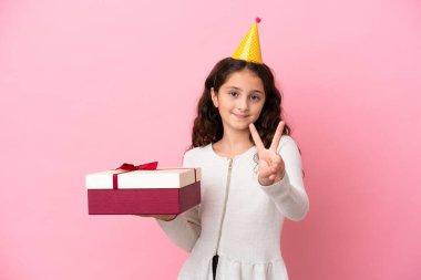 Küçük beyaz kız pembe arka planda izole edilmiş bir hediye tutuyor gülümsüyor ve zafer işareti gösteriyor.