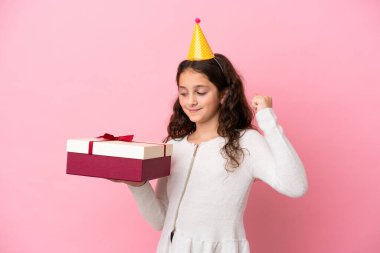 Küçük beyaz kız pembe arka planda izole edilmiş bir hediyeyle zaferi kutluyor.