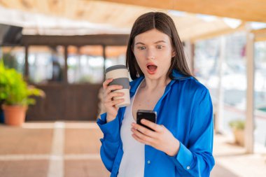 Dışarıda cep telefonu kullanan ve elinde şaşırmış bir ifadeyle kahve tutan genç ve güzel bir kadın.