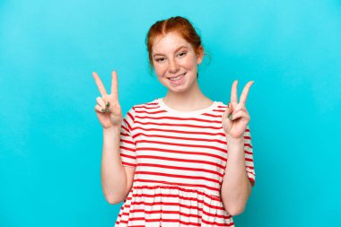 Mavi arka planda izole edilmiş genç kızılımsı kadın iki eliyle zafer işareti gösteriyor.