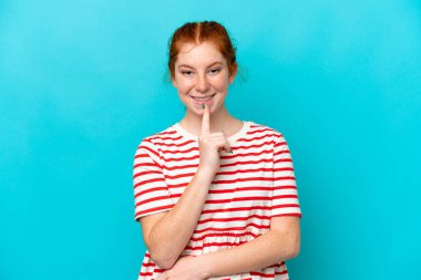 Mavi arka planda izole edilmiş genç kızılımsı bir kadın sessizlik işareti gösteriyor. Parmağını ağzına sokuyor.