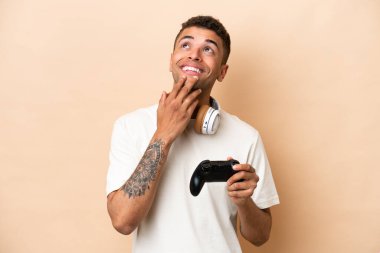Genç Brezilyalı bir adam bej arka planda izole edilmiş bir bilgisayar oyunu kontrolörüyle oynuyor gülümserken yukarı bakıyor