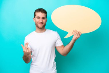 Kafkasyalı genç adam mavi arka planda izole edilmiş, elinde boş bir konuşma balonu var ve geliyor.