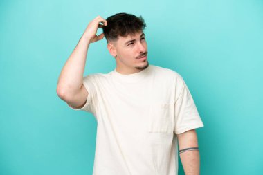 Kafkasyalı yakışıklı genç adam mavi arka planda izole edilmiş, kafası kaşınırken şüpheleri var.