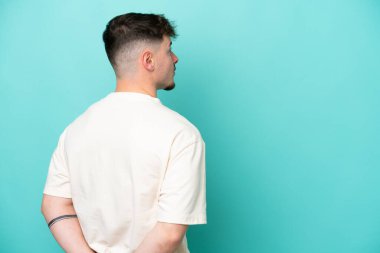 Kafkasyalı yakışıklı genç adam mavi arka planda izole edilmiş ve arkasına bakıyor.