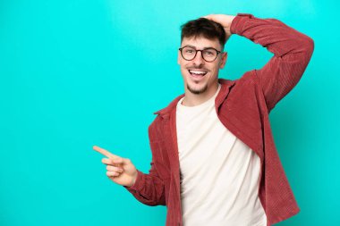 Mavi arka planda izole edilmiş, genç, beyaz, yakışıklı bir adam şaşırmış ve parmağıyla kenarı işaret ediyor.