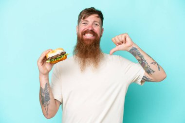 Uzun sakallı kızıl saçlı bir adam mavi arka planda kendini beğenmiş ve gururlu bir burger tutuyor.