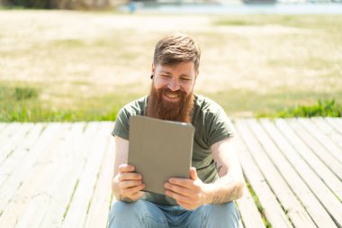 Kızıl saçlı, sakallı, elinde mutlu bir ifadeyle tablet tutan adam.
