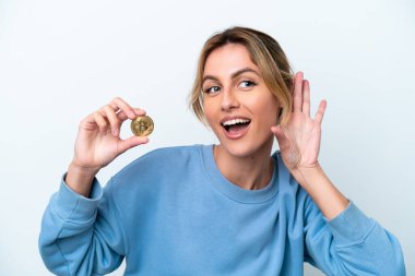Genç Uruguaylı kadın elinde Bitcoin ile beyaz arka planda izole edilmiş bir şekilde elini kulağına koyarak bir şeyler dinliyor.