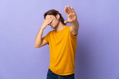Mor arka planda izole edilmiş genç yakışıklı adam dur hareketi yapıyor ve yüzünü kapatıyor.