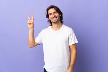 Mor arka planda izole edilmiş genç yakışıklı gülümseyen ve zafer işareti gösteren