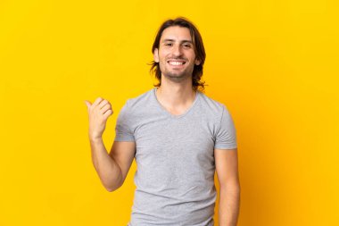 Sarı arka planda izole edilmiş genç yakışıklı bir adam bir ürün sunmak için kenarı işaret ediyor.