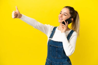 İzole edilmiş sarı arka planda cep telefonu kullanan genç bir kadın işaret veriyor.