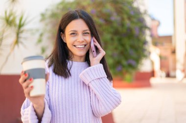 Dışarıda cep telefonu kullanan ve elinde mutlu bir ifadeyle kahve tutan genç bir kadın.