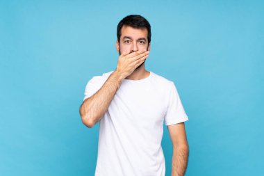 İzole edilmiş mavi arka planda sakallı genç bir adam elleriyle ağzını kapatıyor.