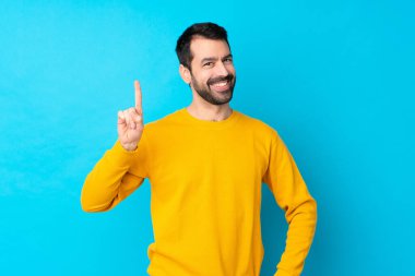İzole edilmiş mavi arka plan üzerinde genç beyaz adam en iyinin işareti olarak parmağını kaldırıyor ve gösteriyor.