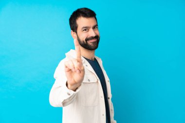 İzole edilmiş mavi arka planda beyaz kadife ceketli genç yakışıklı adam parmağını kaldırıyor ve gösteriyor.